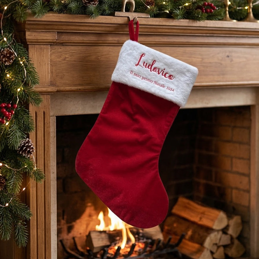 Calza di Natale rossa con bordo bianco, nome e breve scritta stampati, appesa davanti a un camino