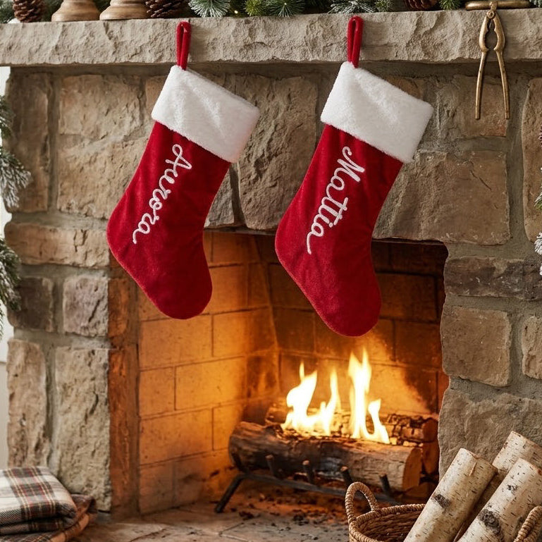 Due calze della Befana rosse con bordo bianco e nomi personalizzati, appese davanti a un camino acceso