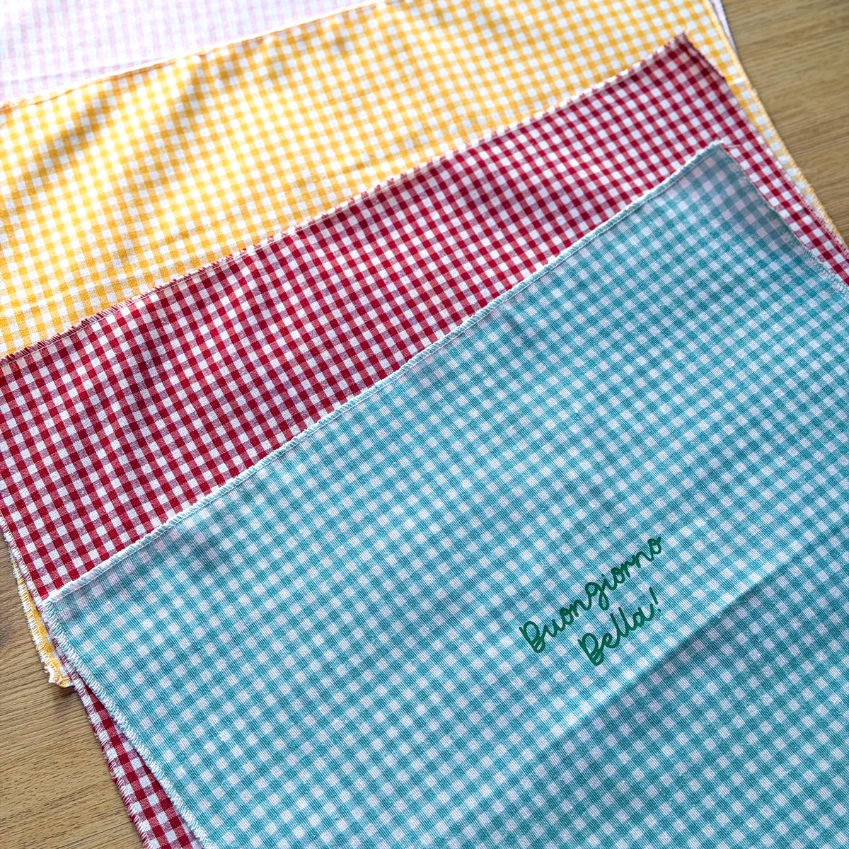 Tovagliette vichy in diversi colori con esempi di scritte personalizzate su un tavolo in legno