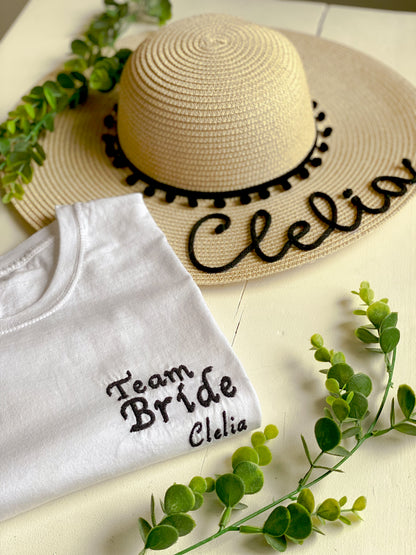tshirt ricamata con scritta "Team Bride" e il nome della ragazza. Accanto c'è un cappello di paglia con scritta del nome e pompon, abbinato alla tshirt