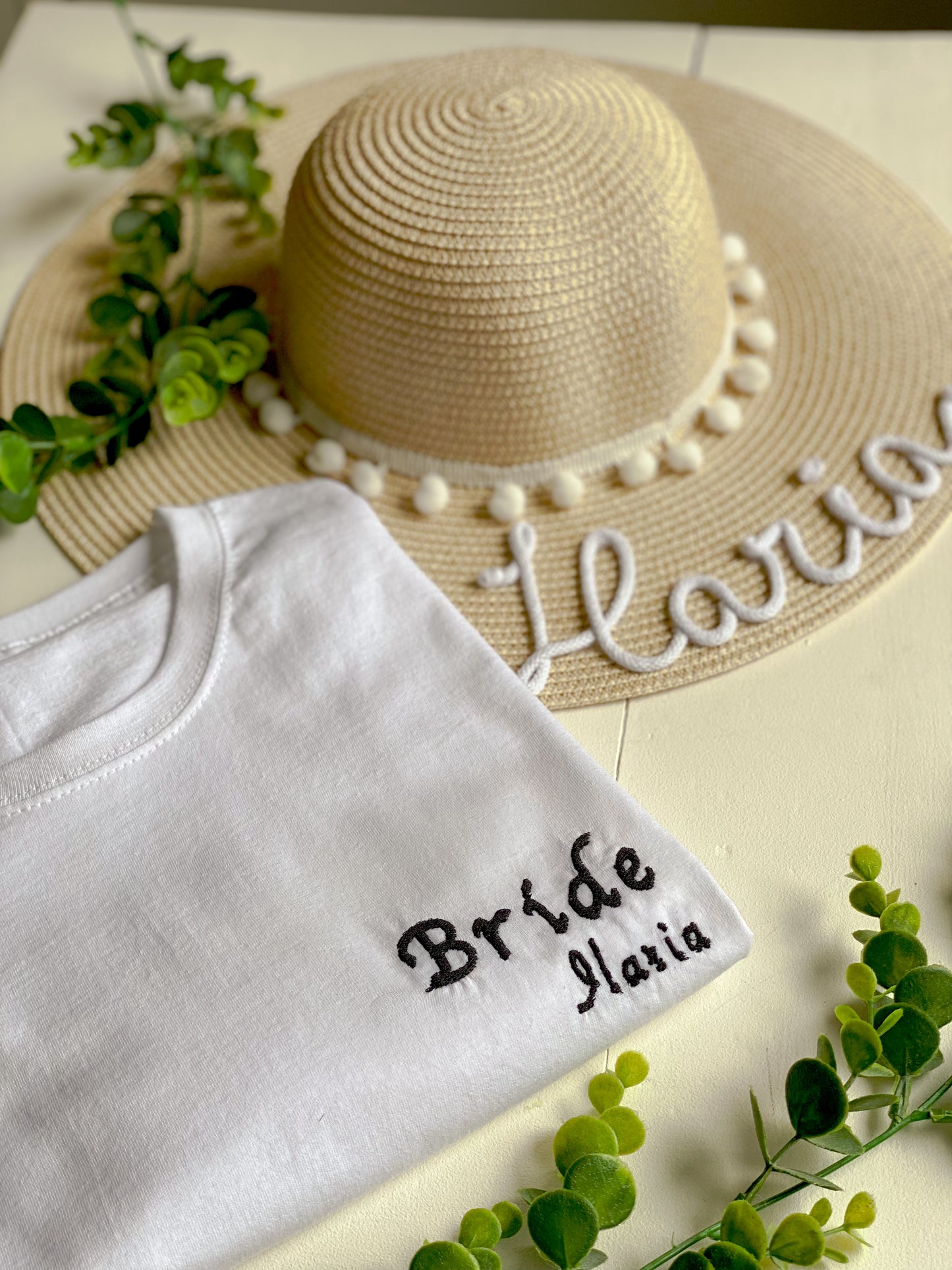 Tshirt ricamata con scritta "Bride" e il nome della ragazza. accanto c'è un cappello di paglia con scritta bianca e pompon bianchi